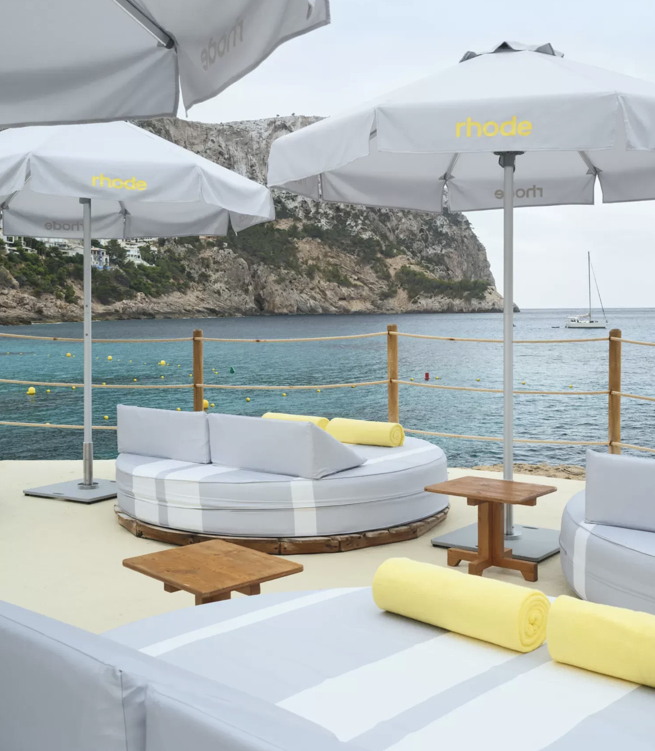Beachside lounge area with white cylindrical seating, yellow towels, small wooden tables, large white umbrellas with yellow text, and ocean view with sailboats and cliffs in the background.