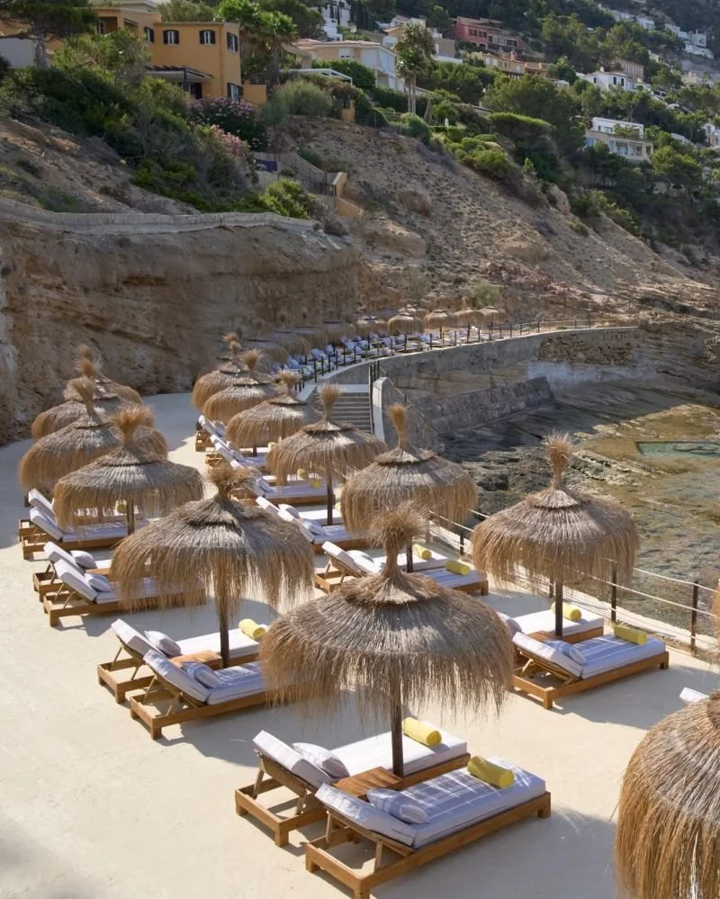 Several empty lounge chairs with yellow towels and white cushions, shaded by straw umbrellas, arranged on a sandy beach with a rocky coastline and hillside houses in the background.