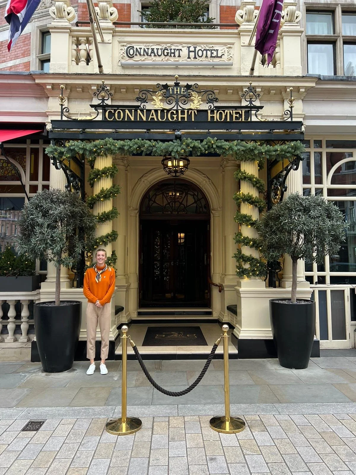 A woman standing in front of the entrance to The Connaught Hotel, decorated with holiday garlands and two potted trees. The hotel has a sign with its name above the door.