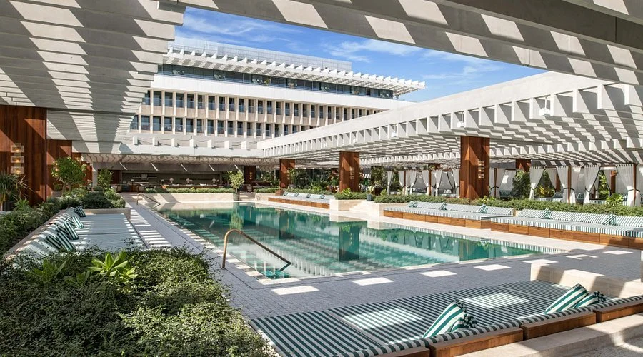 Luxury outdoor pool area with lounge chairs, shaded by modern architectural structures with white beams and wooden supports, surrounded by greenery, in an urban setting.