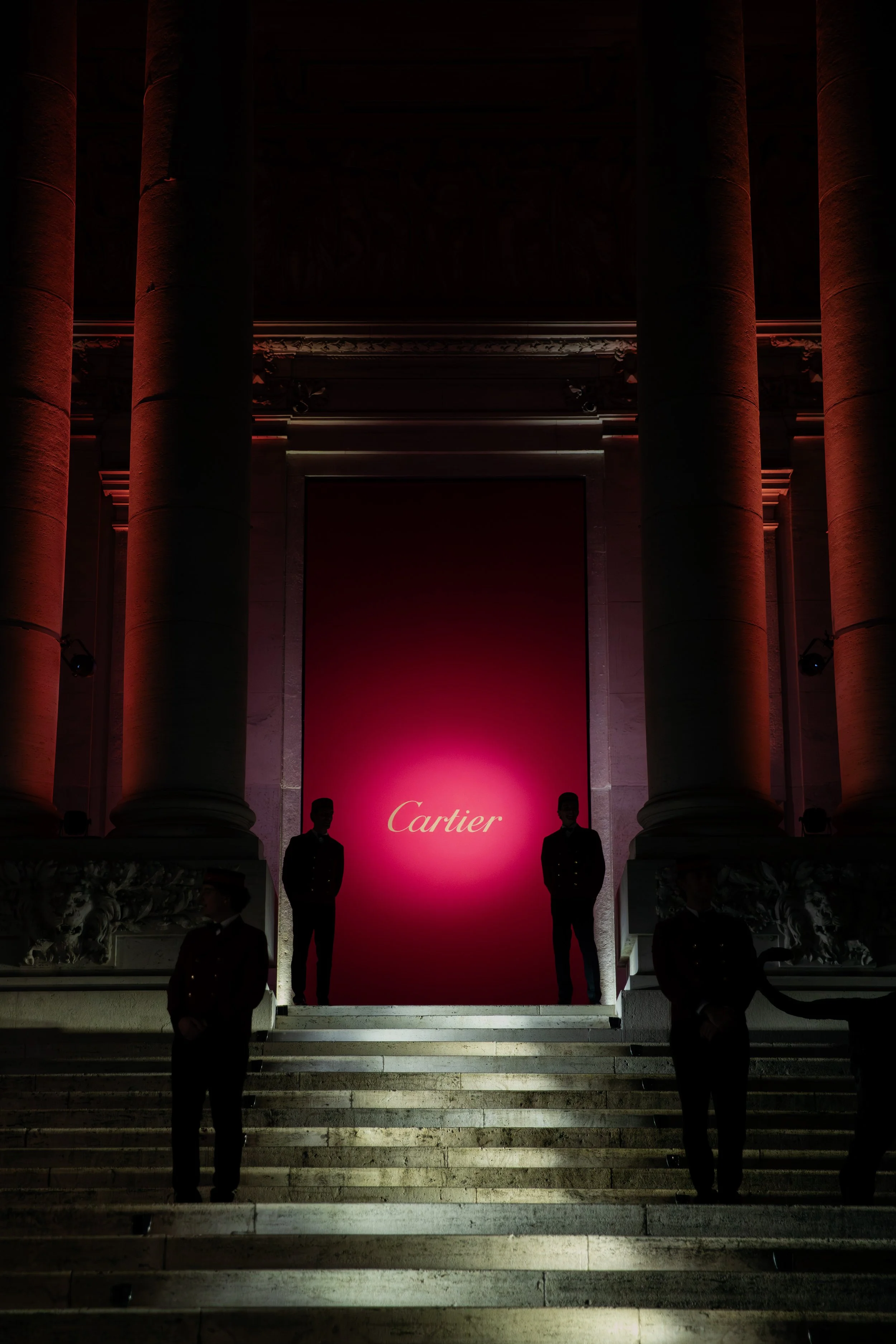 Dark staircase leading up to a large pink backdrop with the Cartier logo, flanked by four security guards in dark uniforms at night.