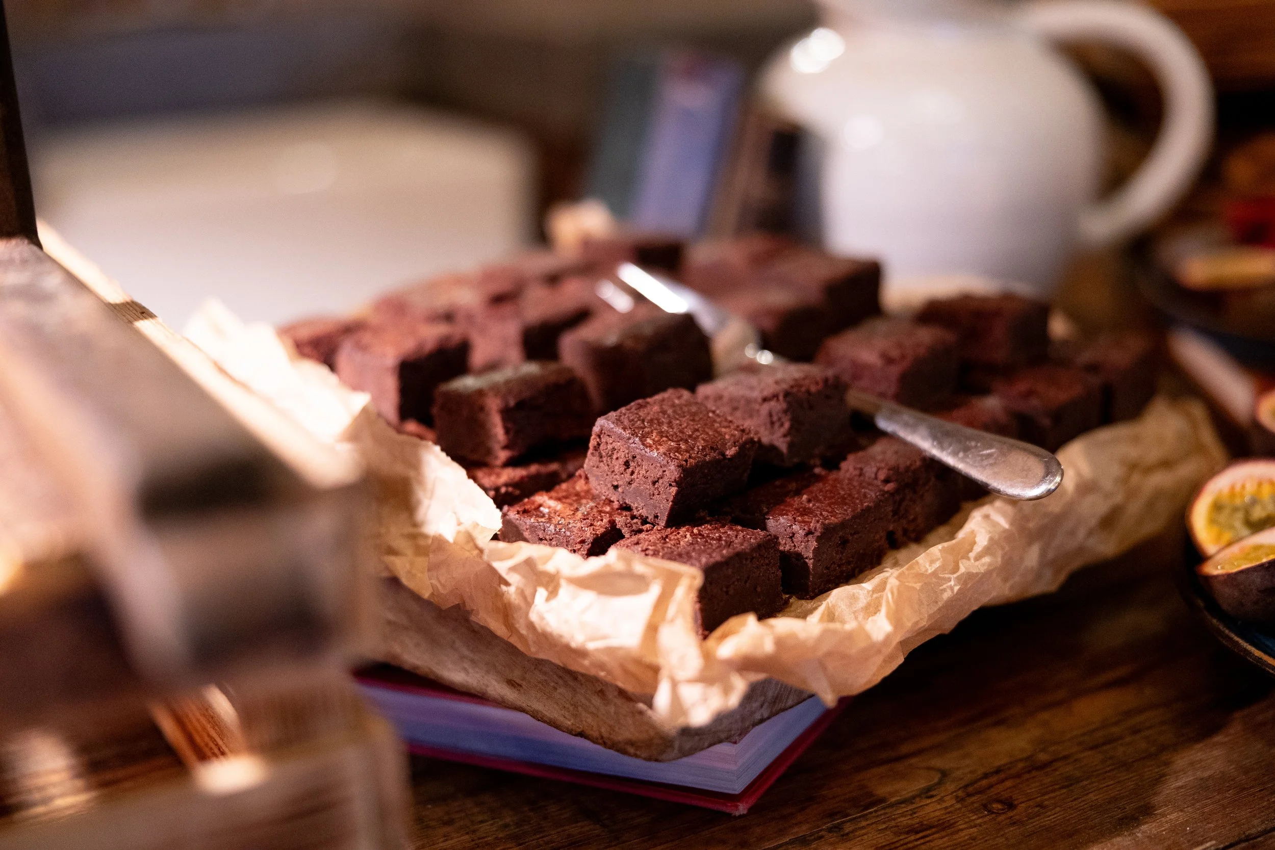 A stack of chocolate brownies on crinkled parchment paper, with a fork resting on them, a white mug, and a phone in the background on a wooden surface.