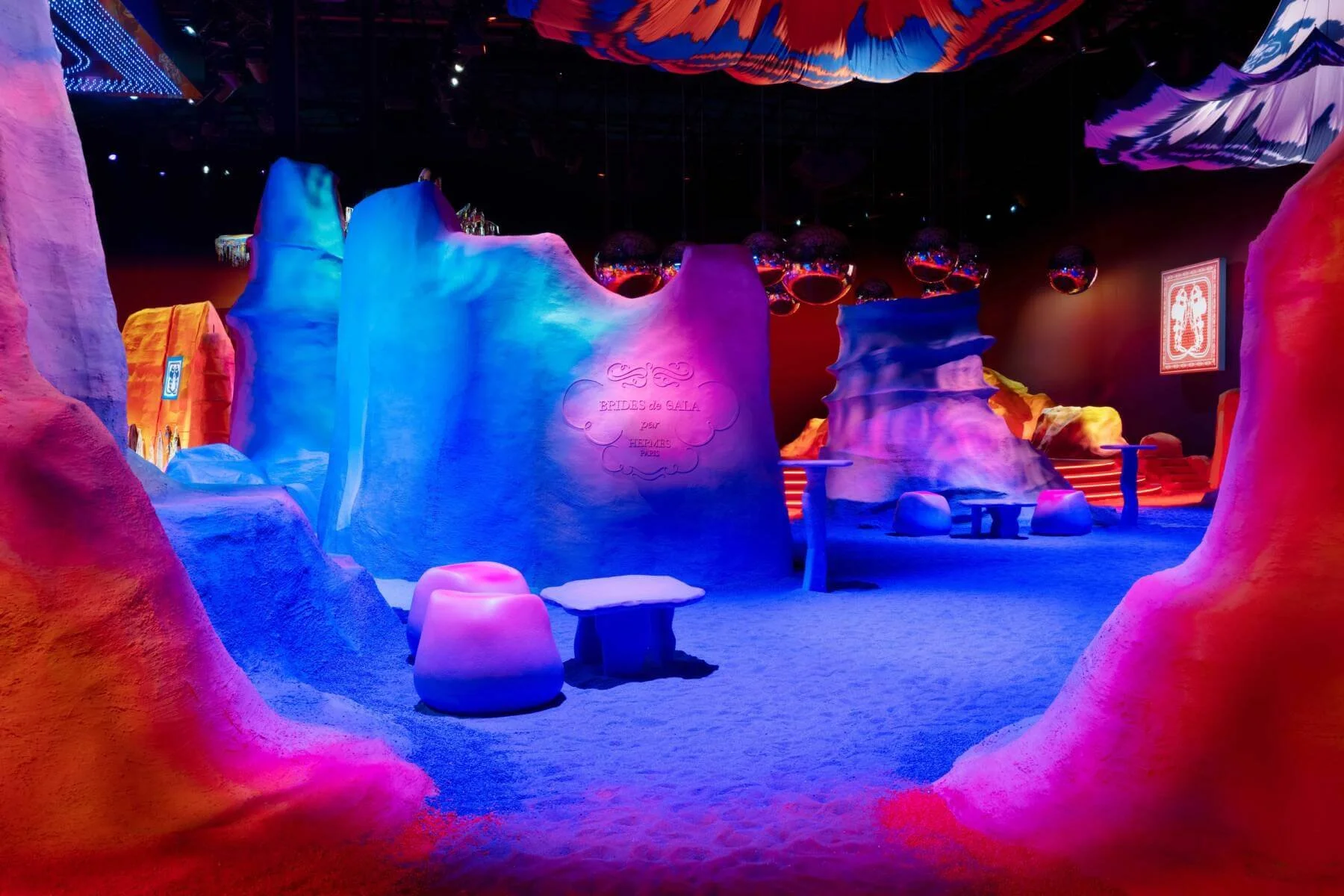 Colorful indoor space with soft, undulating pink, blue, and purple structures resembling abstract rocks or mountains, illuminated with vibrant lighting. There are small tables and stools for seating, and decorative spherical objects hanging from the 