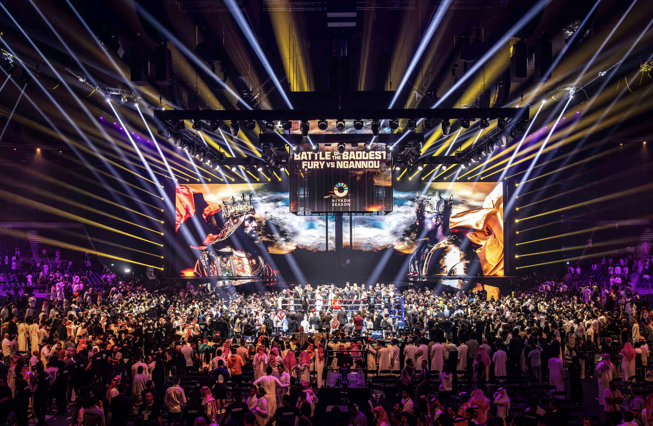 Large indoor arena packed with crowd watching a professional wrestling match. Bright lights and screens display promotional content for a competition titled 'Battle of the Baddest: Fury vs Ngannou', with images of two fighters facing each other. The 