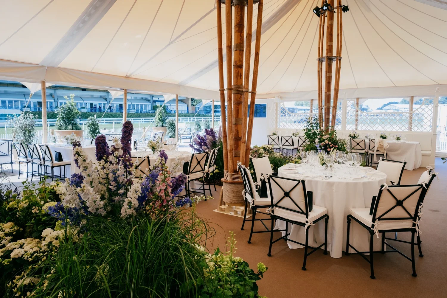 Elegant event space with round tables covered in white tablecloths, set with glassware and silverware, surrounded by black and white chairs, decorated with floral arrangements, inside a tent with a view of a landscaped outdoor area.