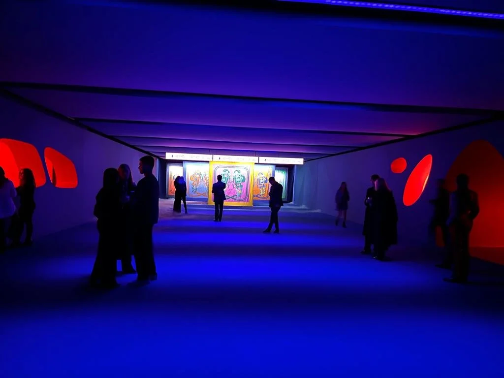 People viewing colorful art displays in a dark, illuminated exhibition space with blue and red lighting.