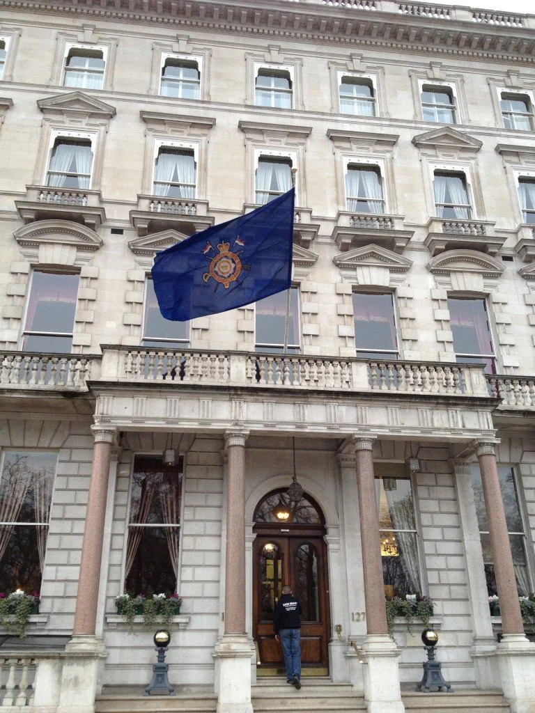 A large, historic building with a flag featuring a crest, two shield-shaped symbols, and gold elements, flying above the entrance. The entrance has three columns, a wooden door, and windows with flower boxes.