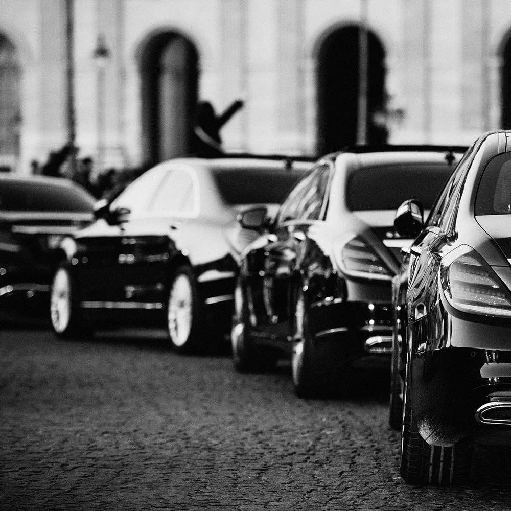 Line of luxury cars parked on a cobblestone street in front of a building with arched windows