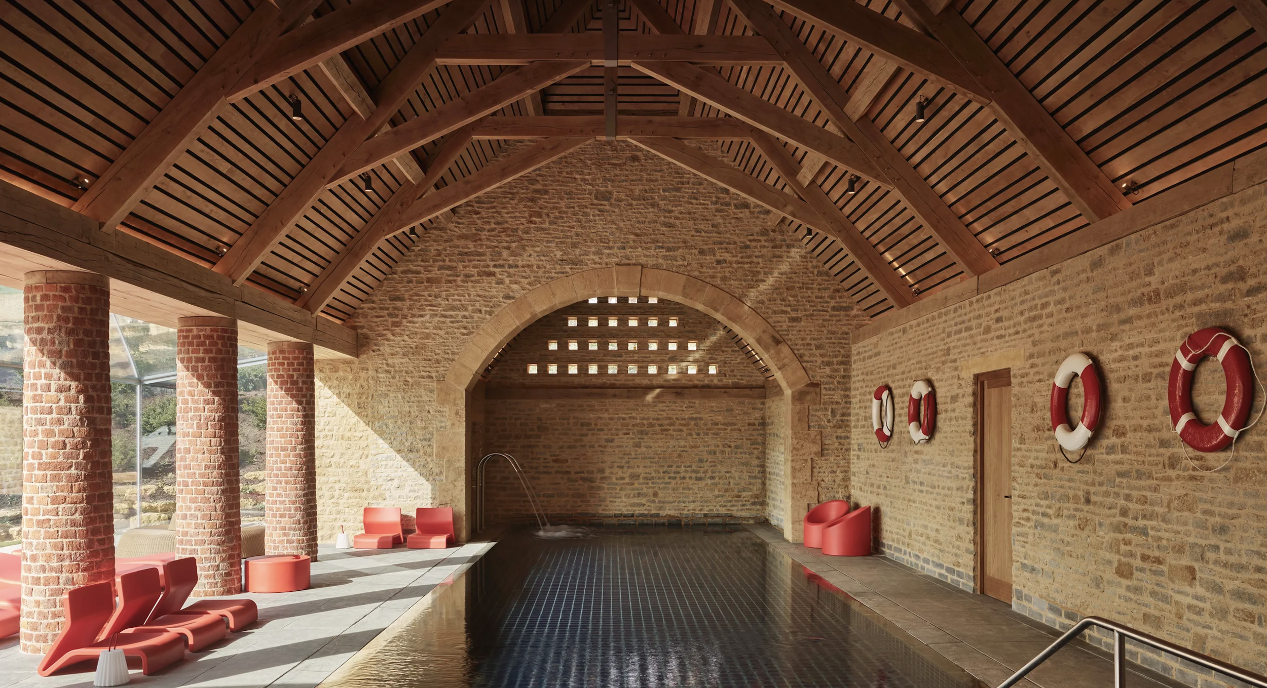 Indoor swimming pool area with brick columns, a brick wall, and a wooden vaulted ceiling. Red lounge chairs and life preservers are mounted on the wall, with sunlight streaming in through large windows.