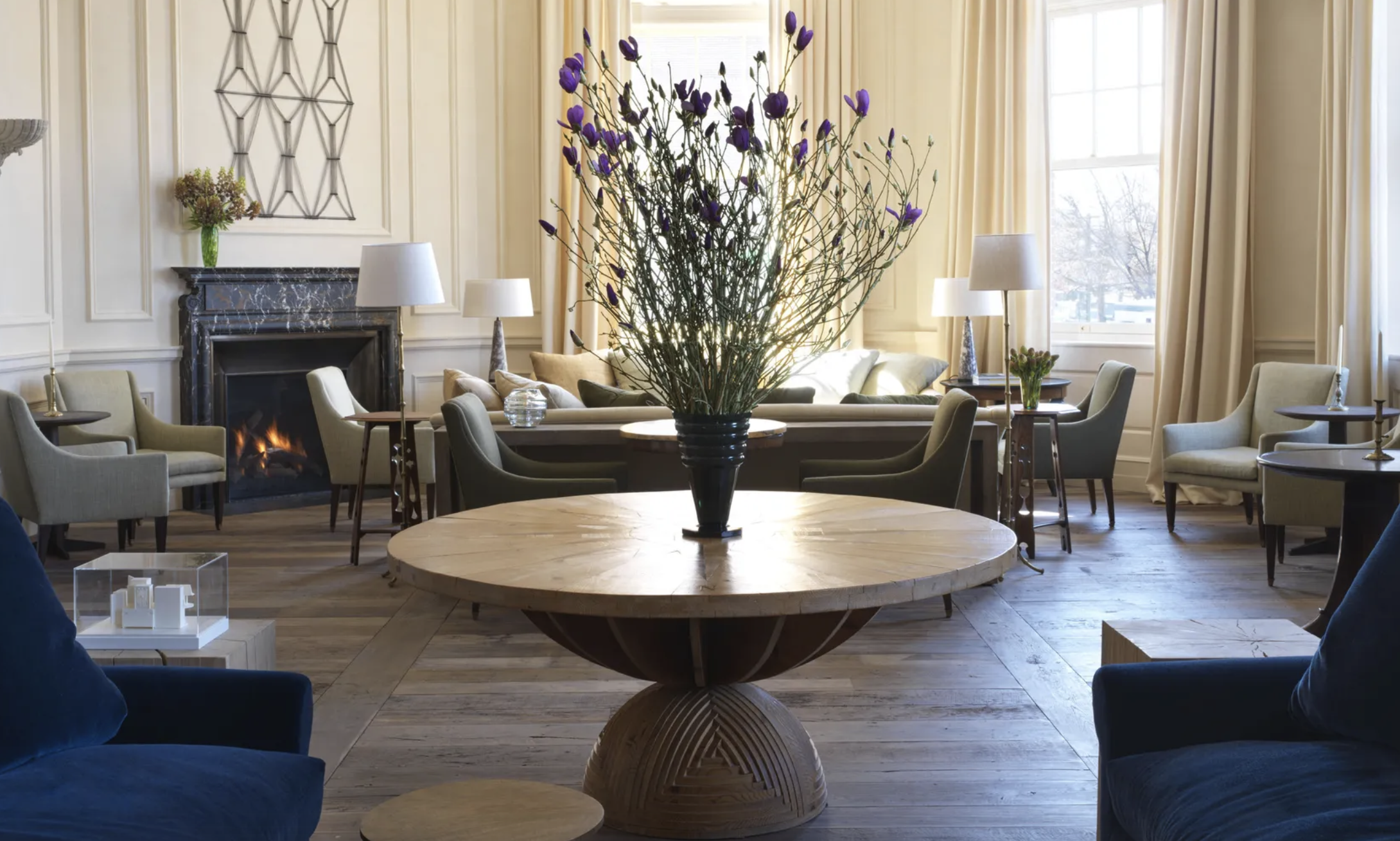 A spacious living room with a large round wooden coffee table, a floral centerpiece with purple flowers, a fireplace with a dark marble mantel, multiple beige armchairs, side tables with lamps, and cream-colored curtains by windows letting in natural