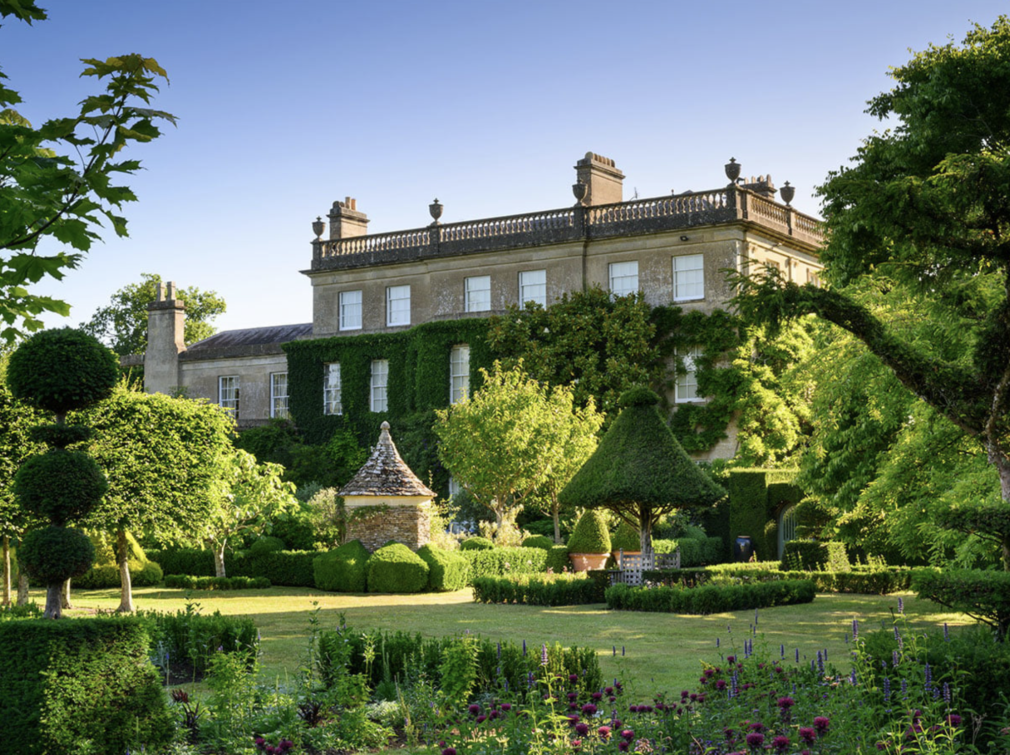 A stately home surrounded by a well-maintained garden with trimmed trees, colorful flowers, and lush greenery, under a clear blue sky.