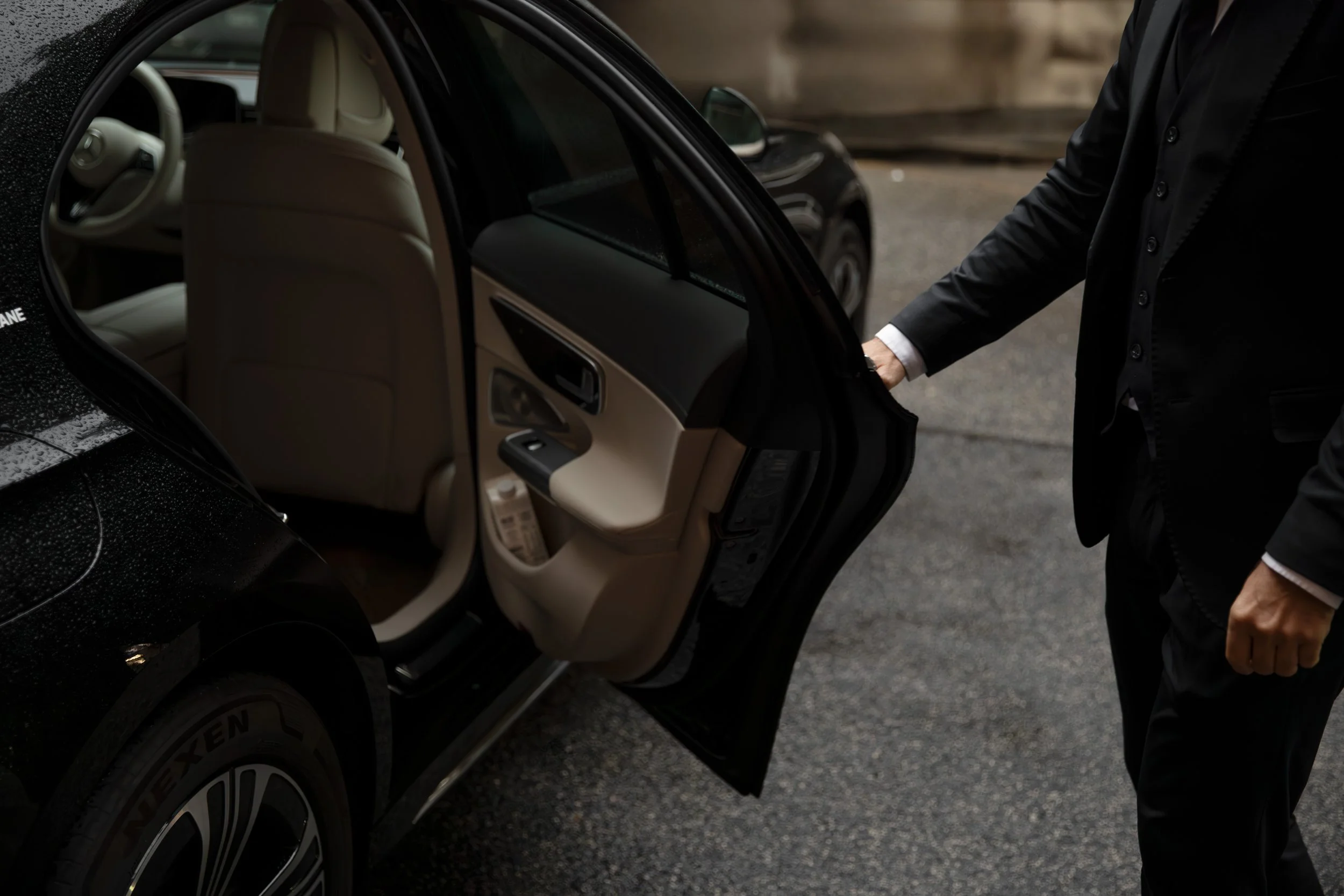 A person in a black suit opening the rear door of a black car, showing the beige interior.