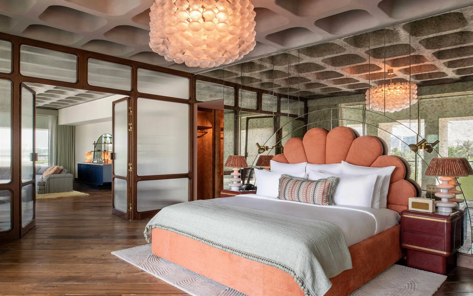 A bedroom with an orange upholstered bed featuring a large, scalloped headboard. There are white pillows and a decorative pillow on the bed, with bedside tables on each side. The room has a mirrored wall behind the bed, wooden floors, and a curved wo