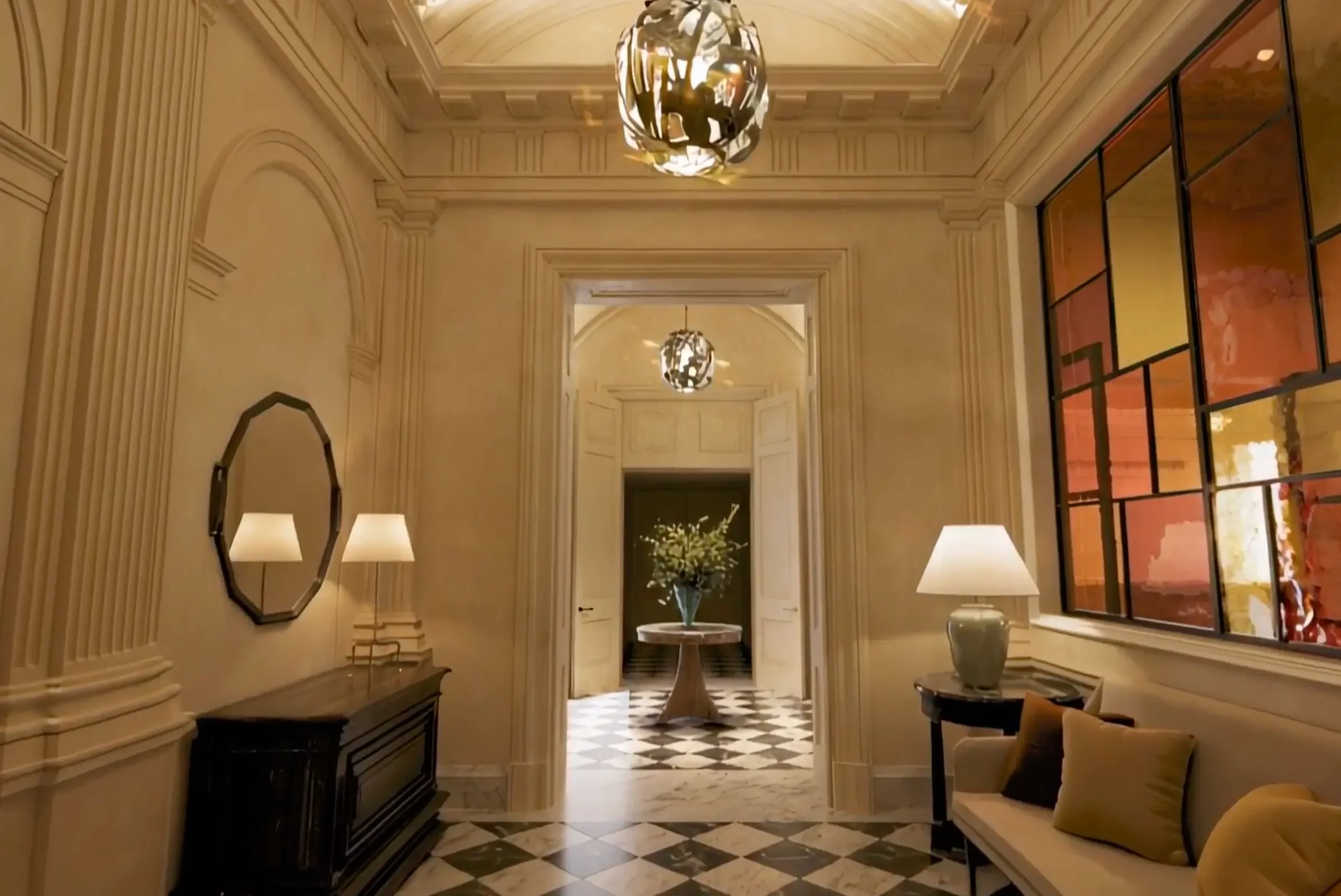 Elegant hotel lobby with cream-colored walls, high ceilings, hanging modern chandeliers, black-and-white checkered marble floor, and decorative furniture, including a table with a floral arrangement in the center and seating areas with cushions.