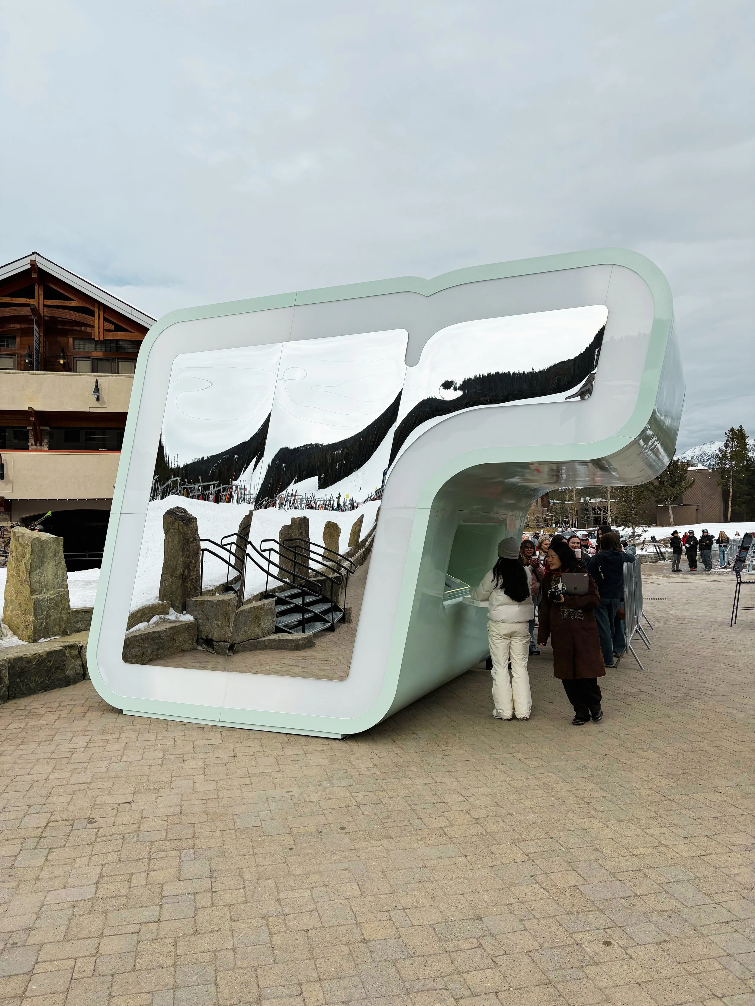 A large, modern mirror sculpture in an outdoor area reflects the surrounding mountains and sky. Several people are gathered around the sculpture, some taking photos, with snow on the ground and mountains in the background.