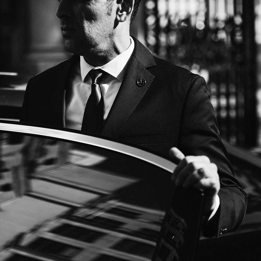 A man in a suit and tie opening the door of a black car in black and white photograph.