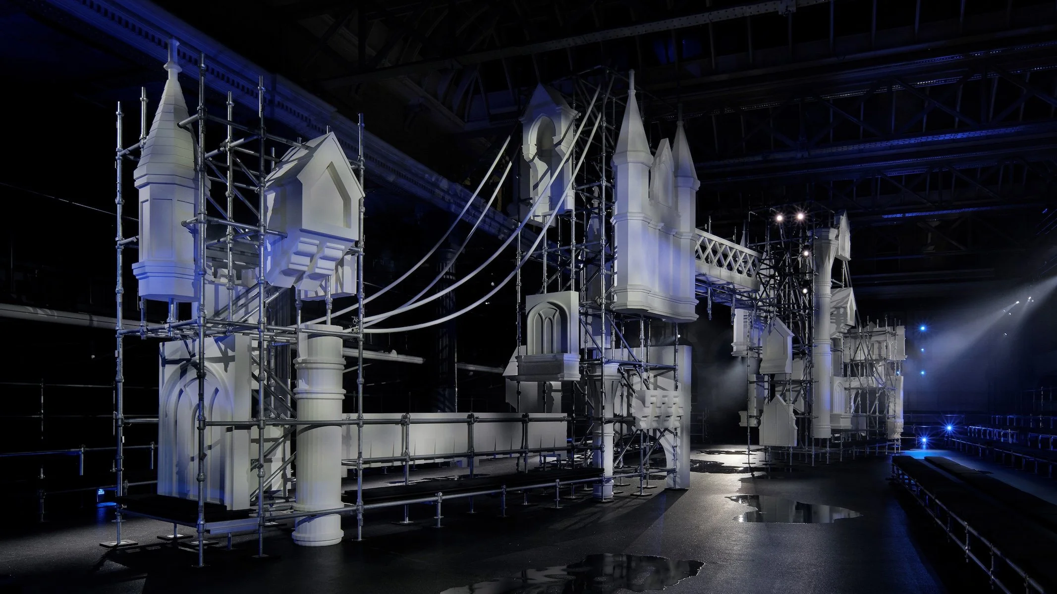 A theatrical set resembling a castle with white turrets under construction, surrounded by scaffolding, in a dimly lit stage environment.