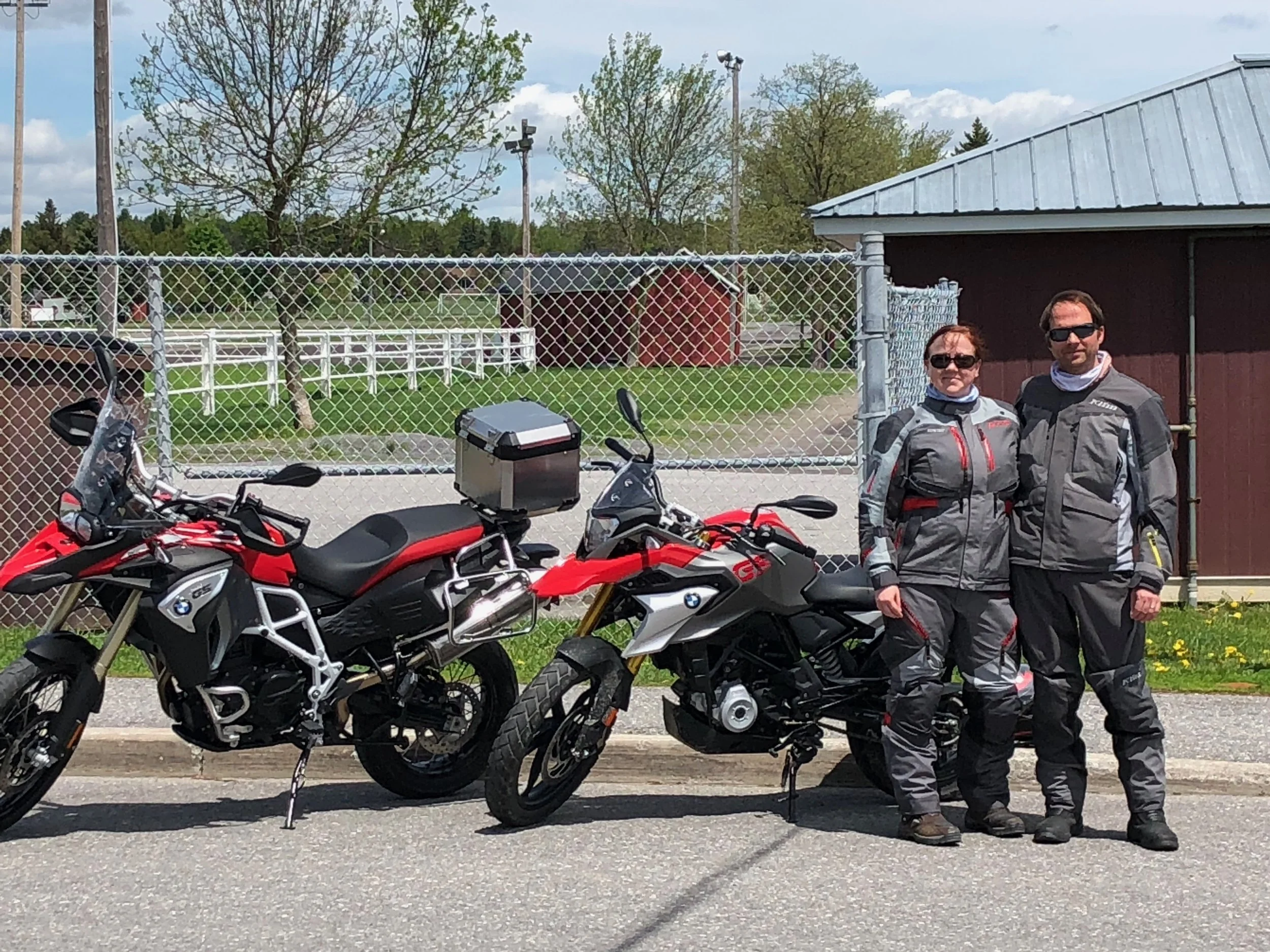 Two motorcyclists wearing riding gear, standing next to their motorcycles on a road with a chain-link fence and a brown building in the background.