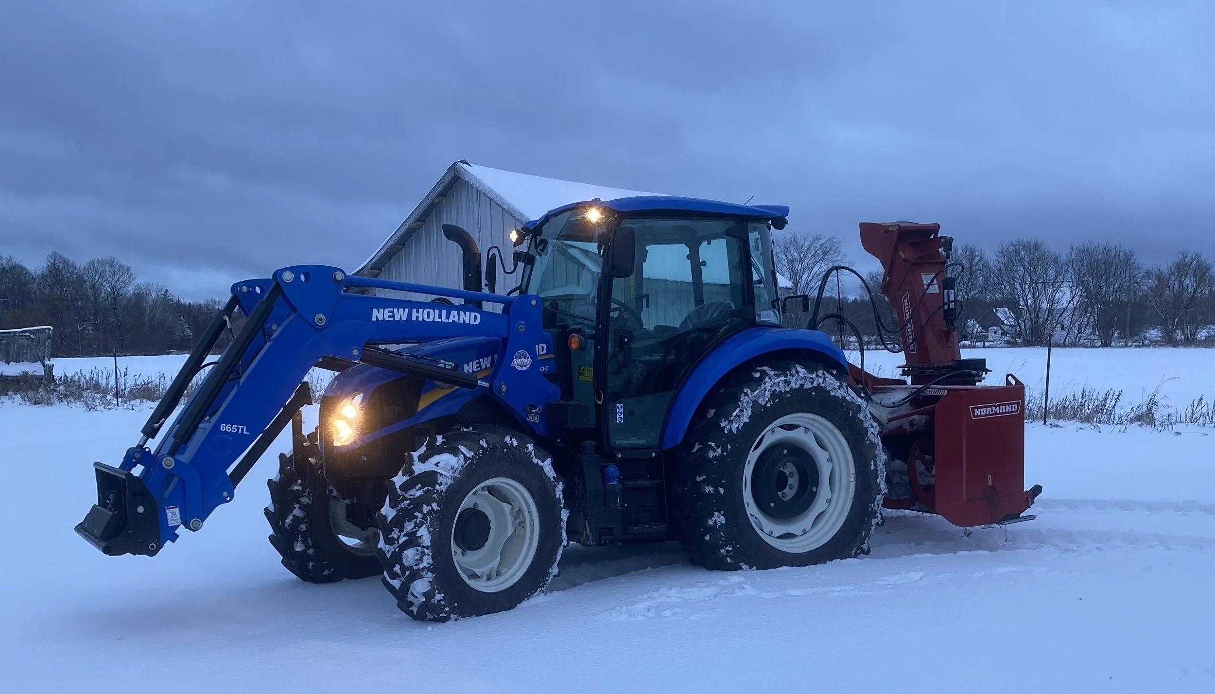 Blue and red New Holland tractor in a snowy field with a barn in the background.