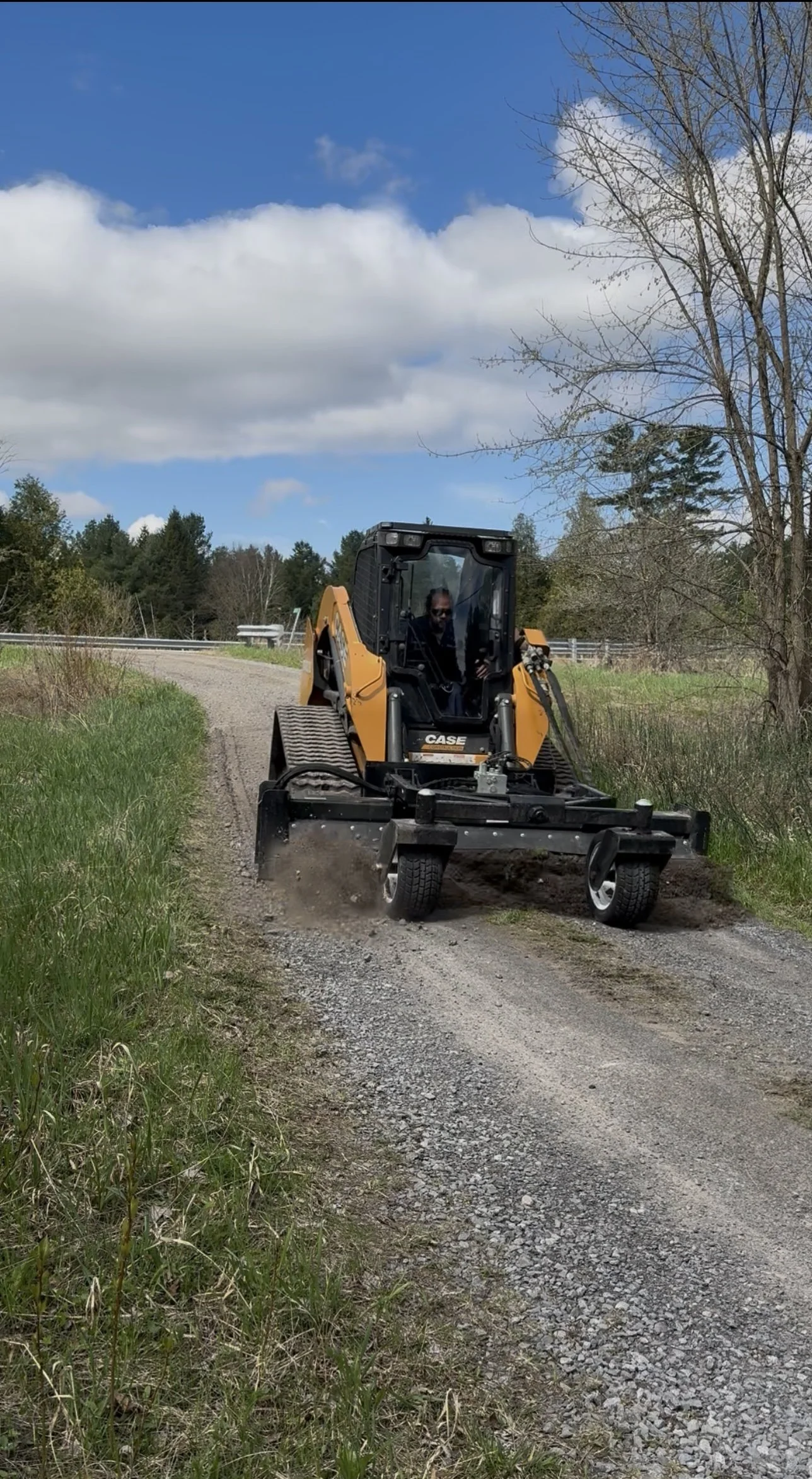 A person operating a yellow CASE compact track loader on a dirt path, clearing and leveling the ground outdoors with a blue sky and trees in the background.