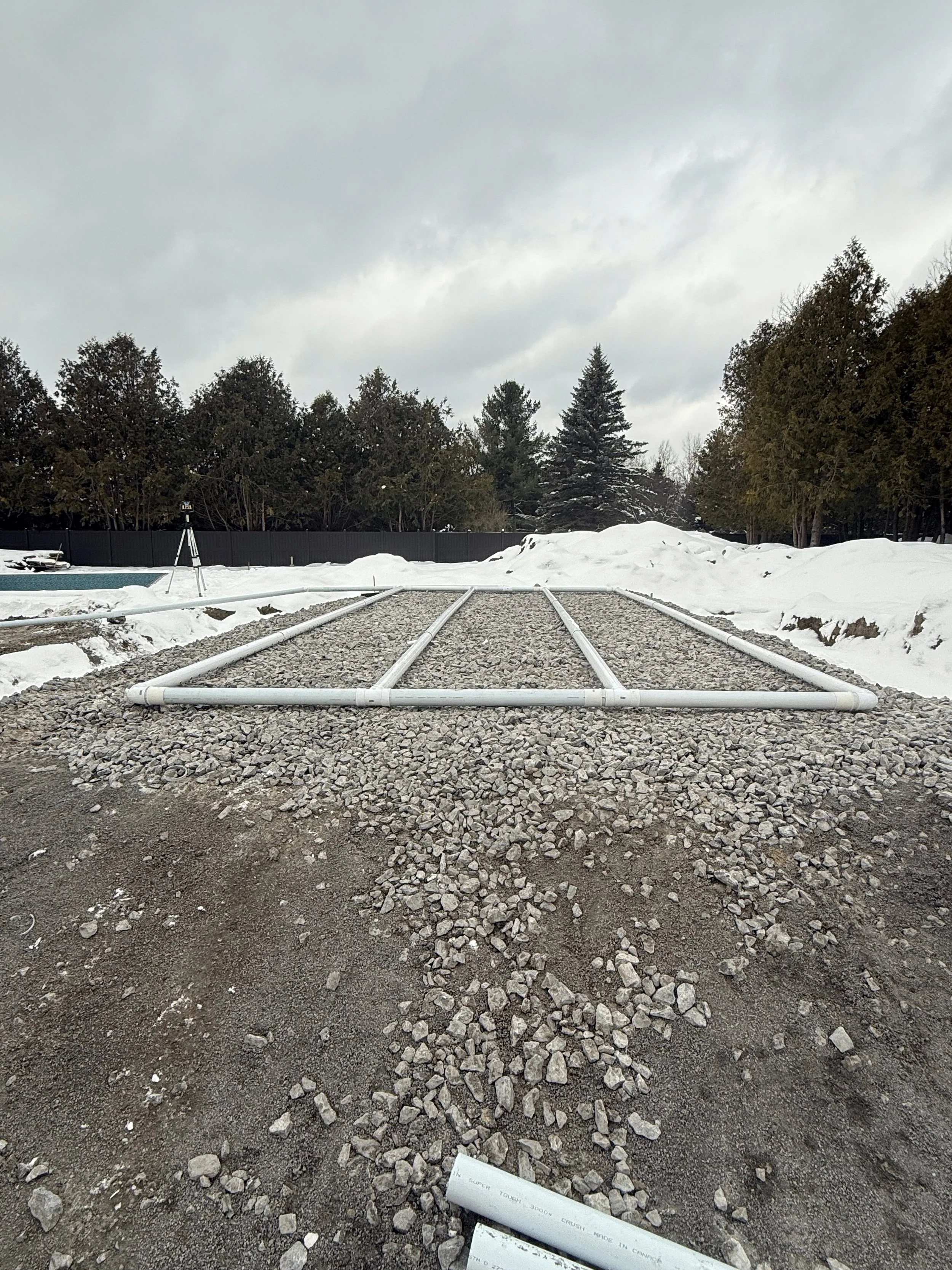 A construction site with a small excavator and a surveyor's tripod with a theodolite, near a barn and silo in a rural area during overcast weather.