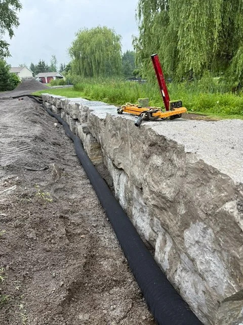 Construction site with a stone wall, black drainage pipe underneath, and construction tools on top of the wall, in a semi-rural area with trees and houses in the background.