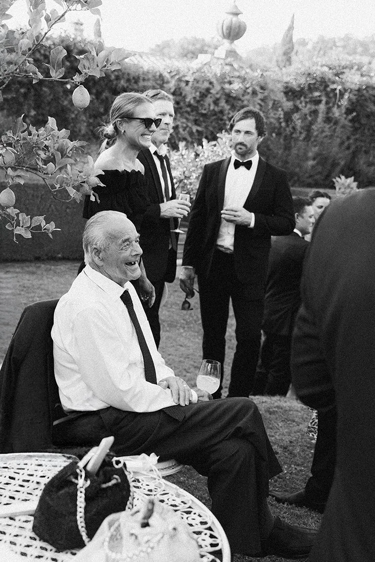 People at a social gathering outdoors, with a man sitting and smiling holding a drink, others standing and chatting, dressed in formal attire.