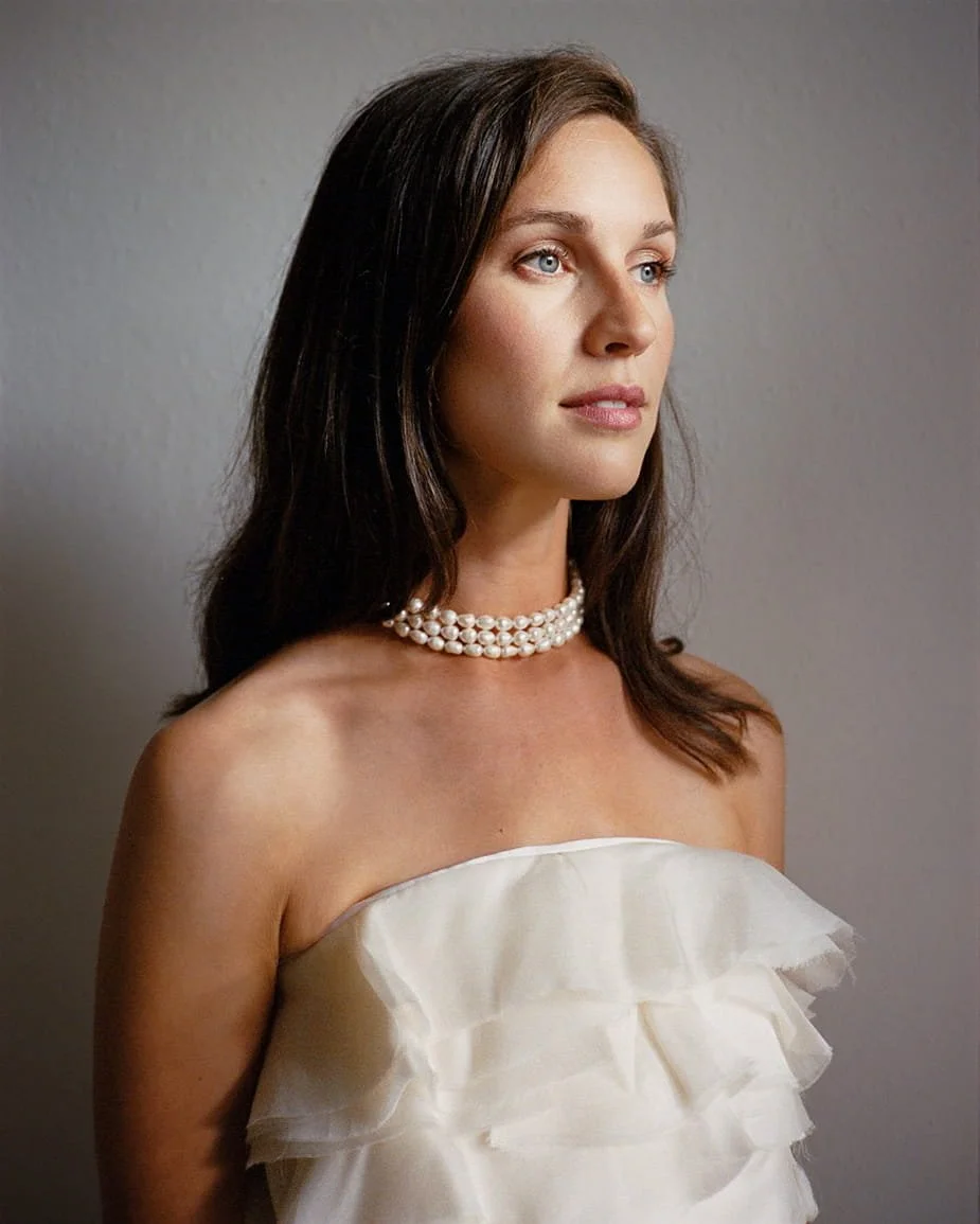 A woman with long brown hair and blue eyes wearing a strapless white ruffled dress and a three-strand pearl necklace, standing against a gray background looking off to the side.