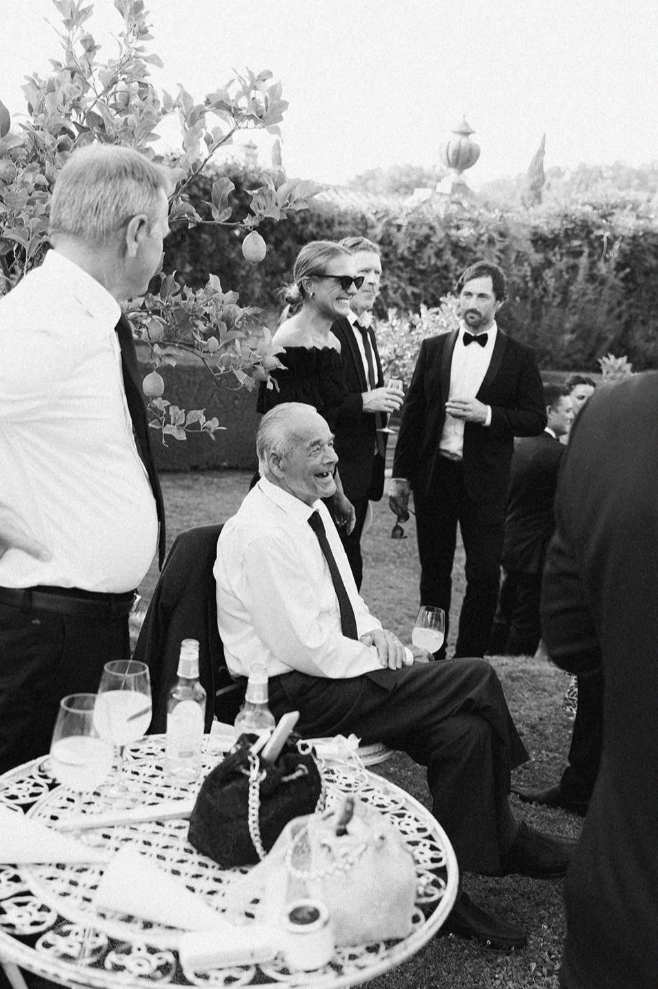 People at an outdoor social gathering, wearing formal attire, with one man sitting and others standing around him, glasses in hand, and a table with drinks in the foreground.
