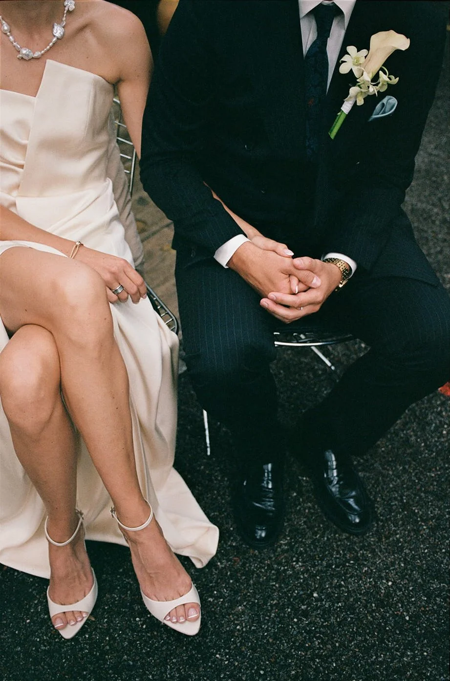 Close-up of a woman in a white dress and a man in a dark suit, holding hands at a formal event or wedding, both sitting on black chairs.