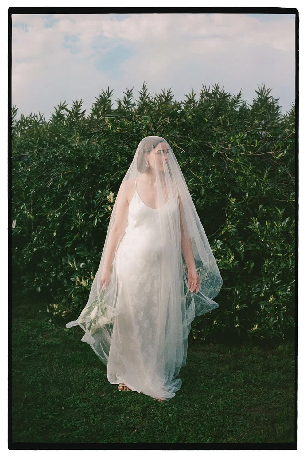 A bride in a white dress with a veil holding white flowers stands outdoors in front of green bushes under a cloudy sky.