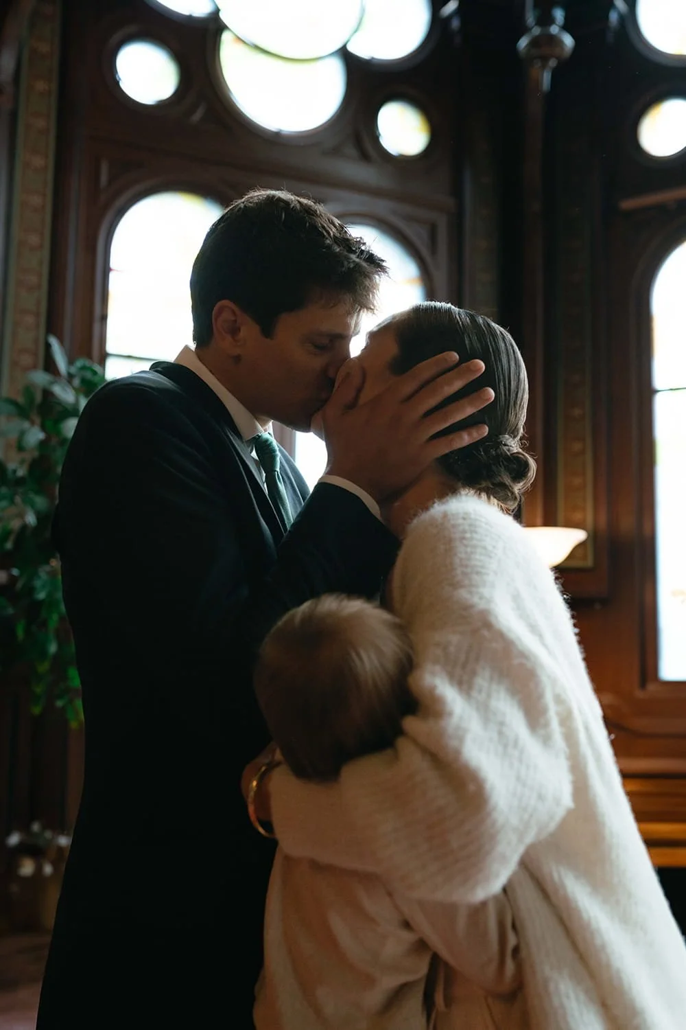 A man and woman are sharing a kiss indoors, with the woman holding a small child in her arms. The background shows large, decorative windows and wooden paneling.
