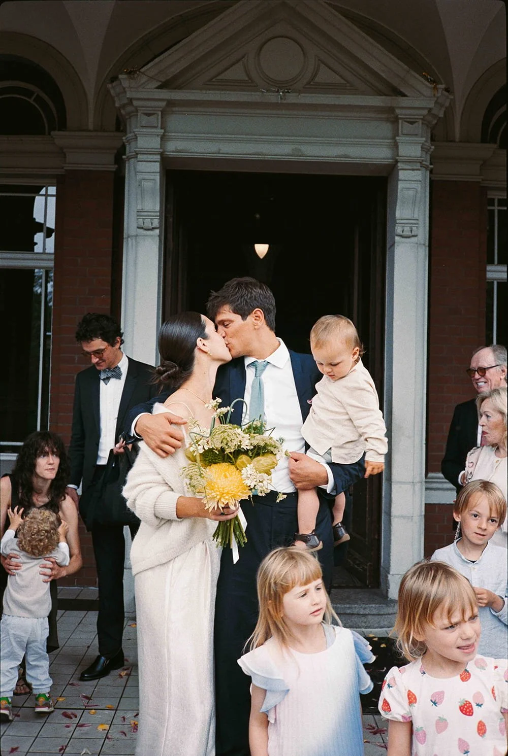 A couple kissing during their wedding celebration, surrounded by children and guests outside a building.