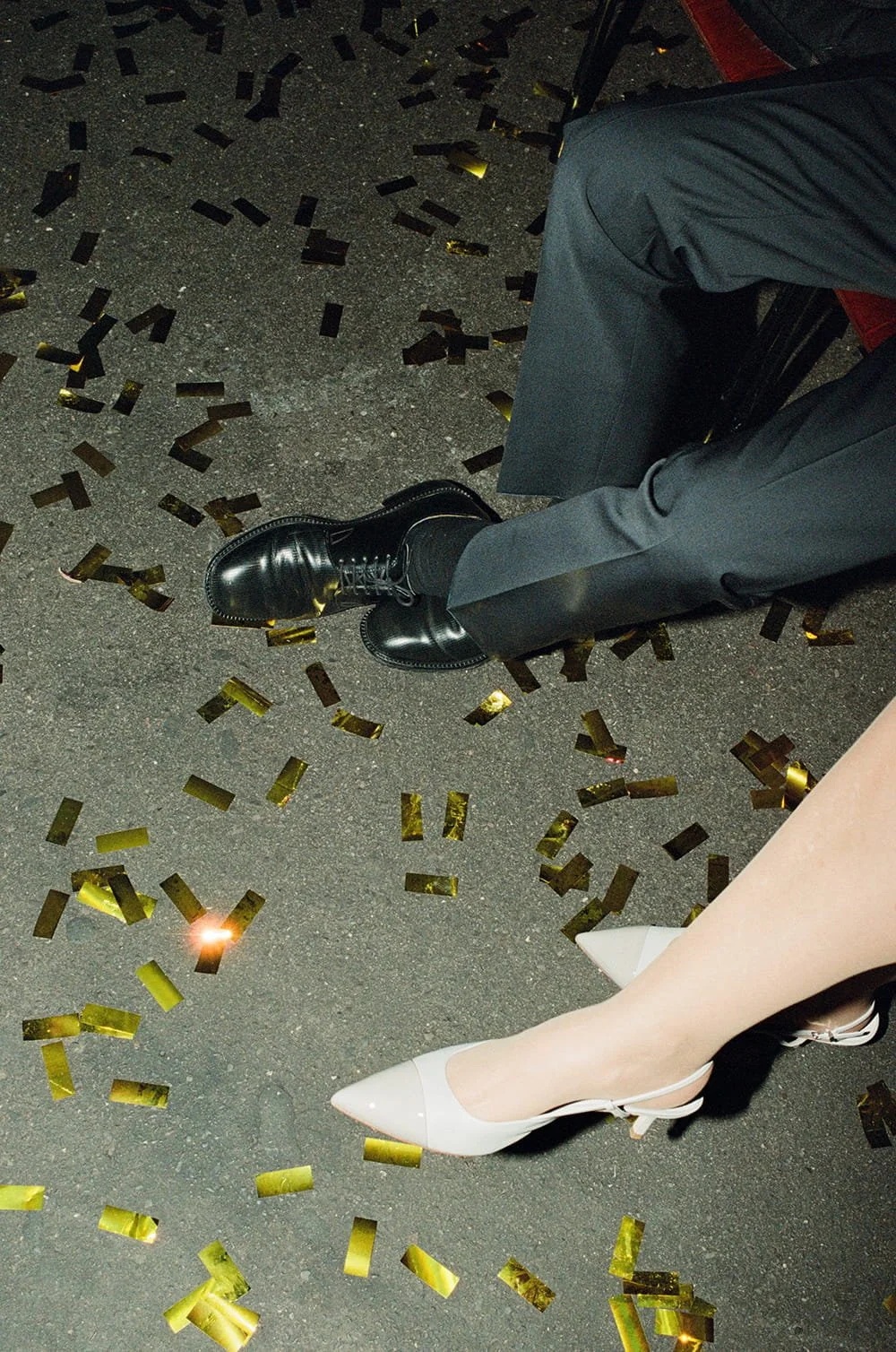 Close-up of a person's legs and feet at a celebration, with golden confetti on the ground, wearing shiny black boots and white high heels.
