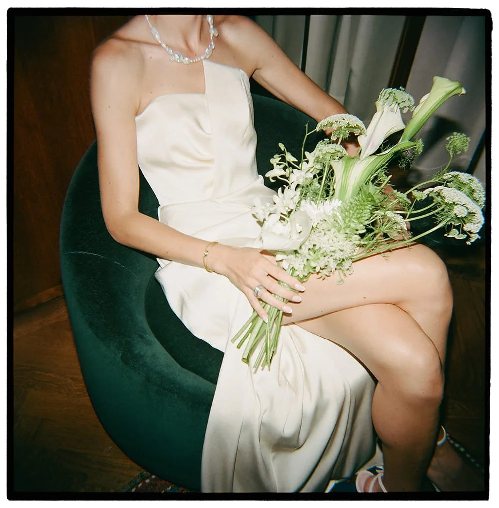 A woman in a strapless white dress, adorned with a pearl necklace and bracelet, sits on a dark green velvet chair holding a large bouquet of white and green flowers.