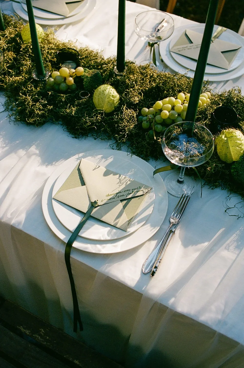 A table setting with a white tablecloth, a white plate, a folded napkin with writing, and silverware. The centerpiece features green candles, grapes, moss, and decorative gourds. There are wine glasses and an envelope or menu on the napkin.