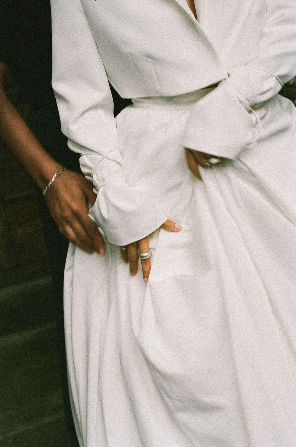 Close-up of a woman wearing a white dress, with hands adorned with rings and a bracelet, holding the fabric of her dress.