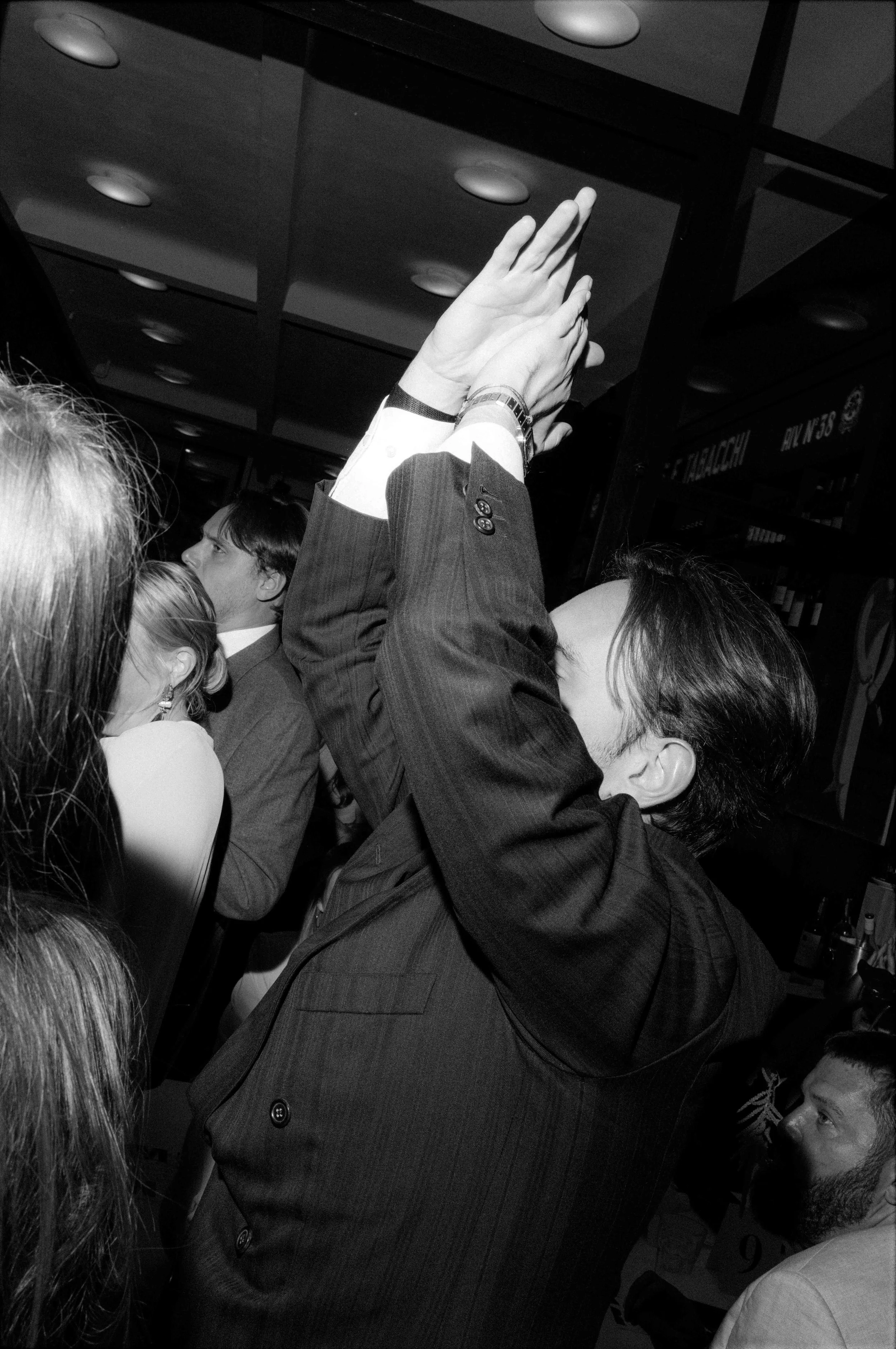 A man in a suit with short dark hair is clapping with his hands raised together at what appears to be a social gathering or event, surrounded by other people in a dimly lit indoor setting.