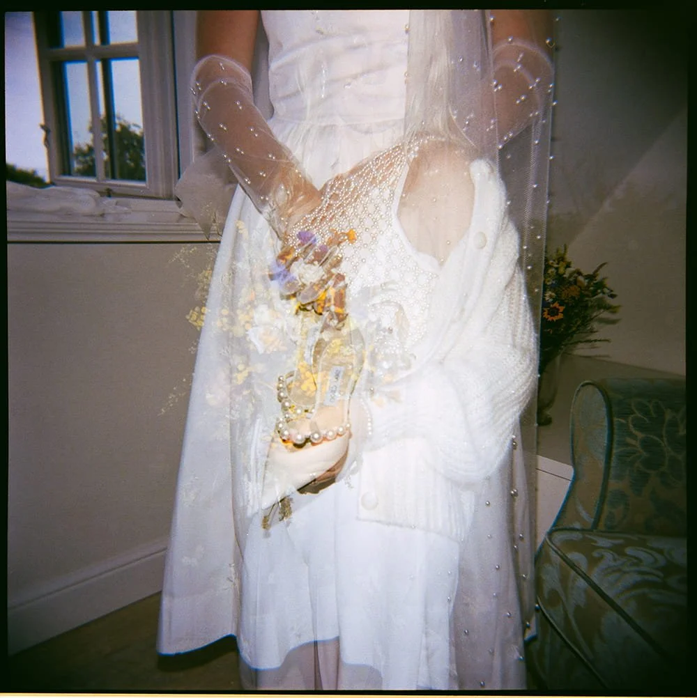 Close-up of a bride in a white dress and lace gloves holding a bouquet of flowers, standing near a window with a view of trees outside.