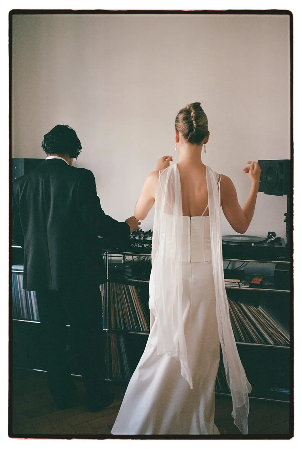 A DJ and a woman in a white dress dancing in a room with vinyl records and audio equipment.
