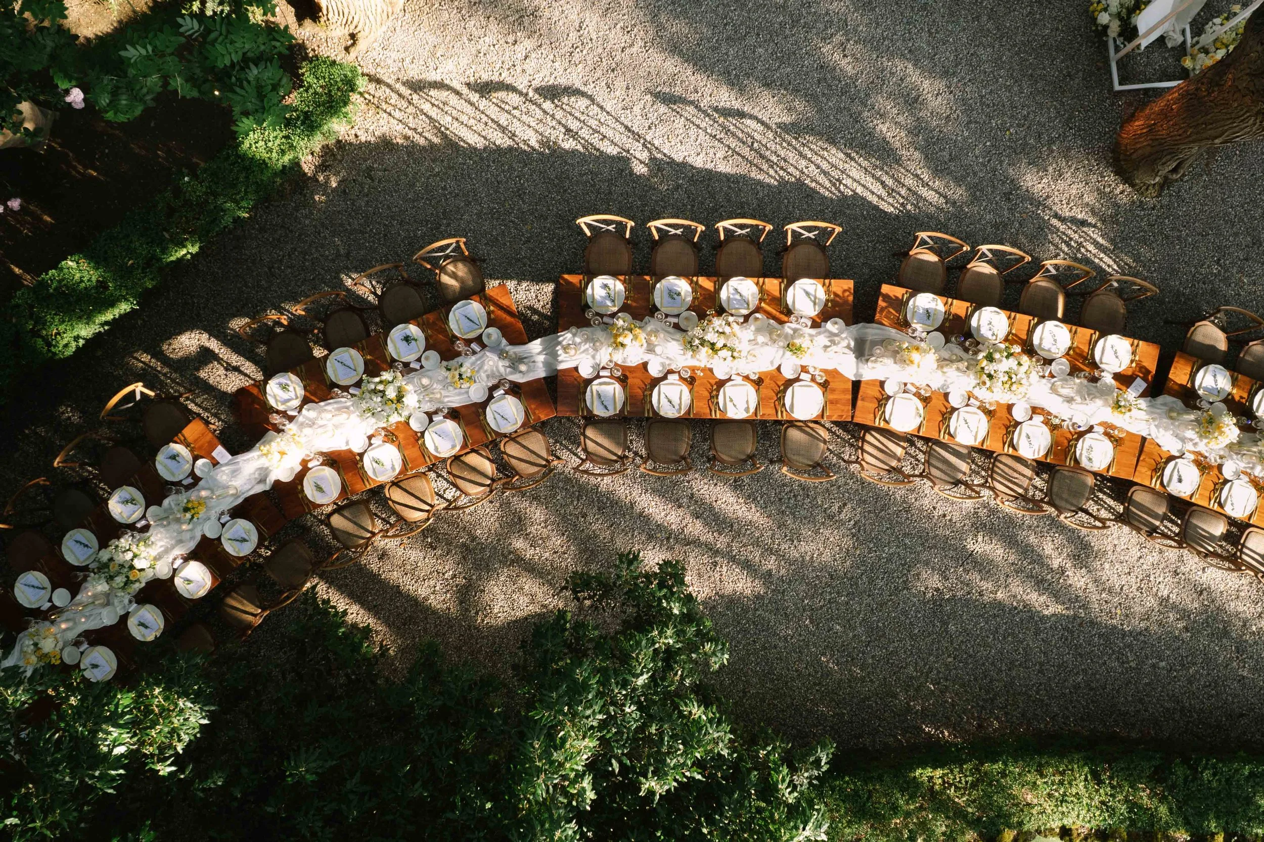 Overhead view of outdoor wedding reception tables decorated with white flowers and tableware, arranged in a curved formation on a gravel surface surrounded by greenery and trees, with shadows cast by nearby objects.