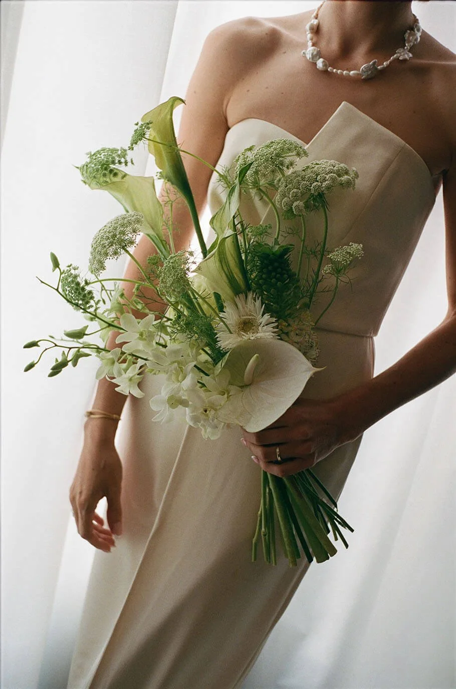 A woman in a strapless white dress holding a bouquet of white and green flowers, including calla lilies, daisies, and greenery, with a pearl necklace and a ring.