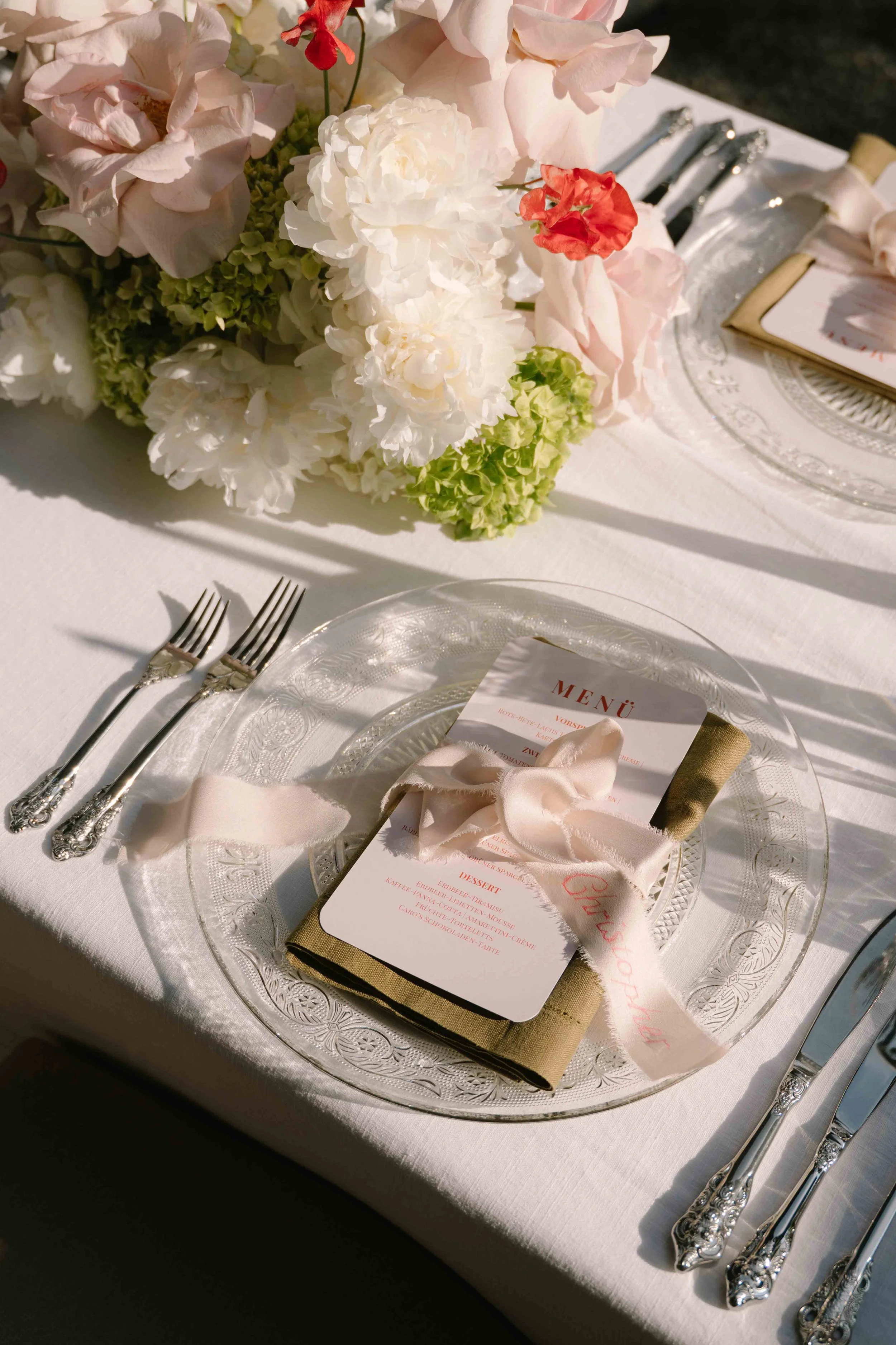 Elegant table setting with floral centerpiece, glass charger plate, menu card tied with a ribbon, and silver cutlery on a white tablecloth.