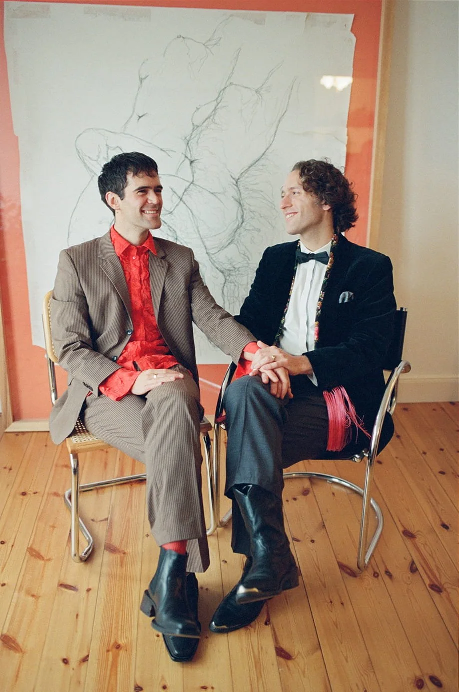 Two men sitting on chairs holding hands and smiling at each other in a room with wooden flooring and a large sketch of a person on the wall behind them. One is wearing a striped suit with a red shirt, the other a black velvet jacket with a bow tie.
