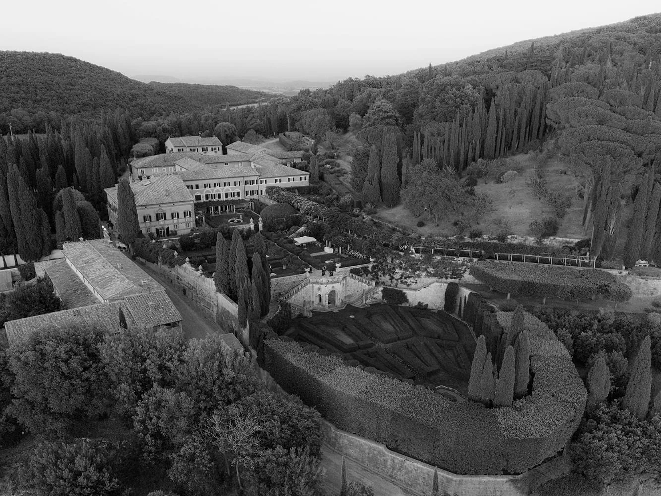 A black and white aerial view of a large estate surrounded by trees and hills, featuring multiple buildings, a landscaped garden, and terraced garden beds.