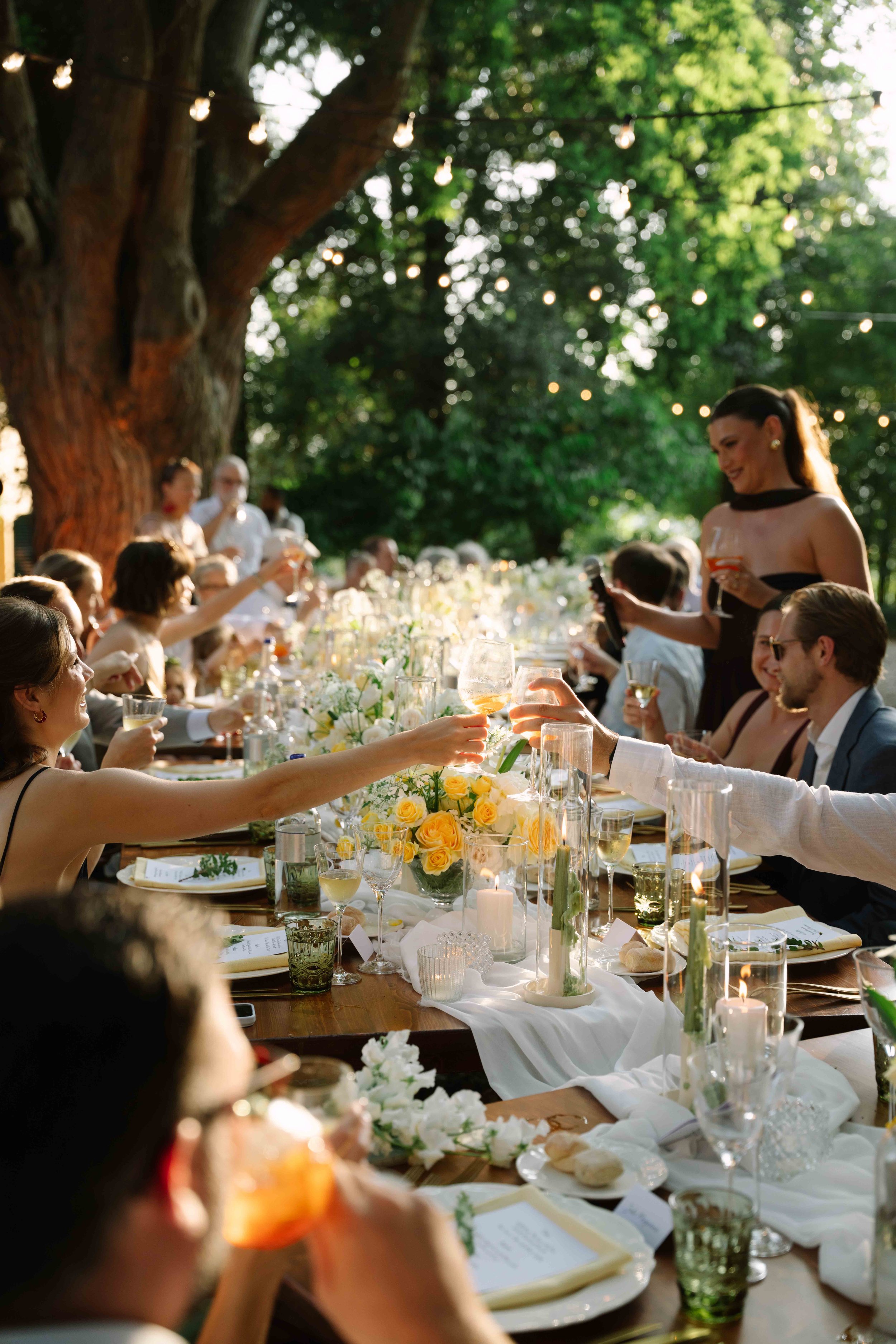 Guests celebrating at an outdoor wedding reception, raising glasses in toast, with a long table decorated with flowers, candles, and elegant tableware under string lights and large trees.