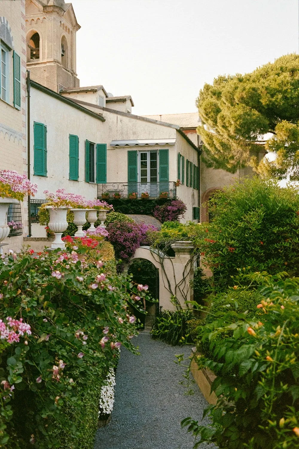 A narrow pathway lined with lush green bushes and colorful flowers leads to a white house with green shutters and a small balcony, surrounded by trees and a stone wall.
