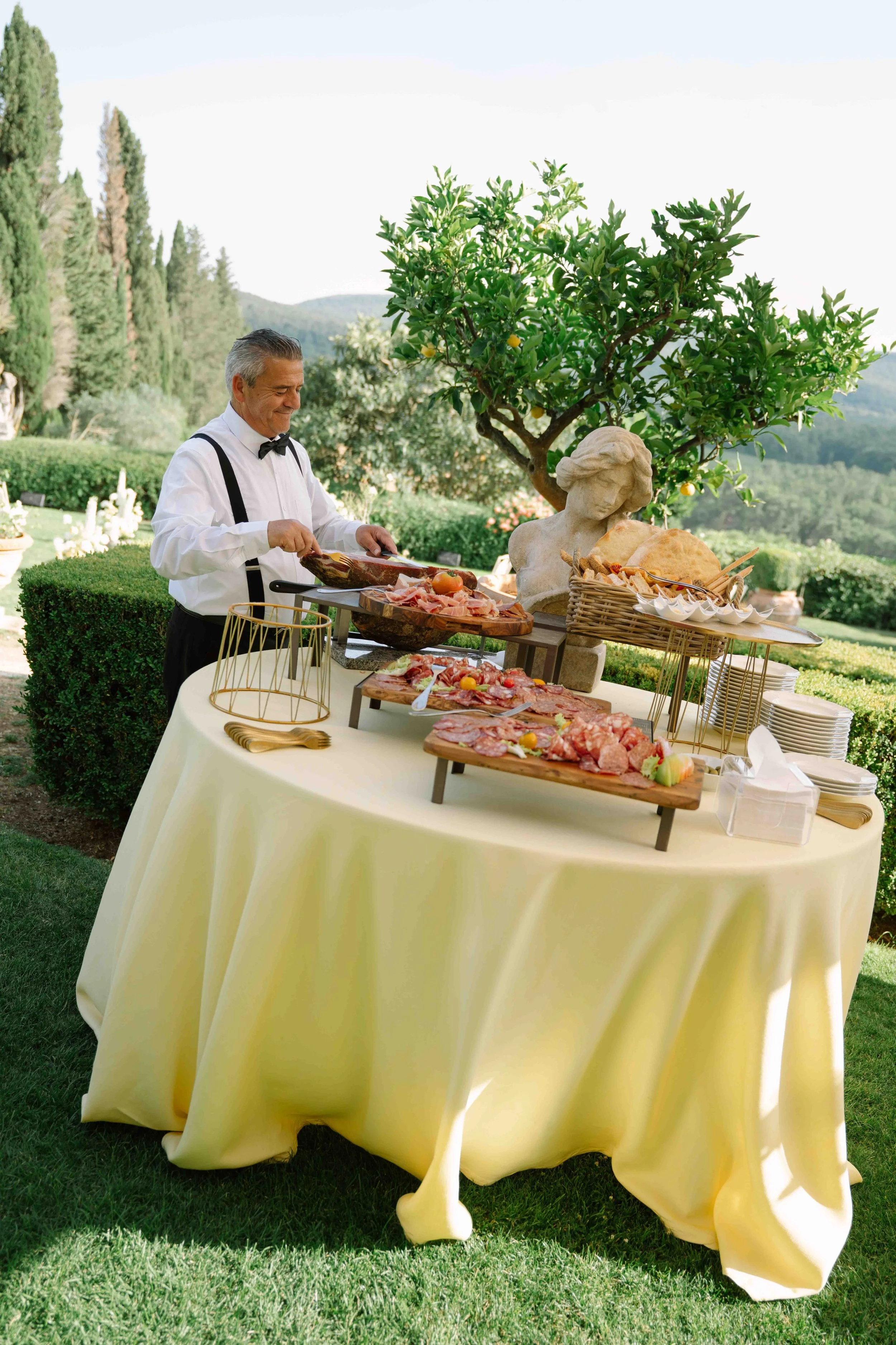 A man in a tuxedo with suspenders serves himself at a buffet table outside, with trees and hills in the background. The table has a yellow tablecloth and is set with various types of cooked meats, bread, and small plates.