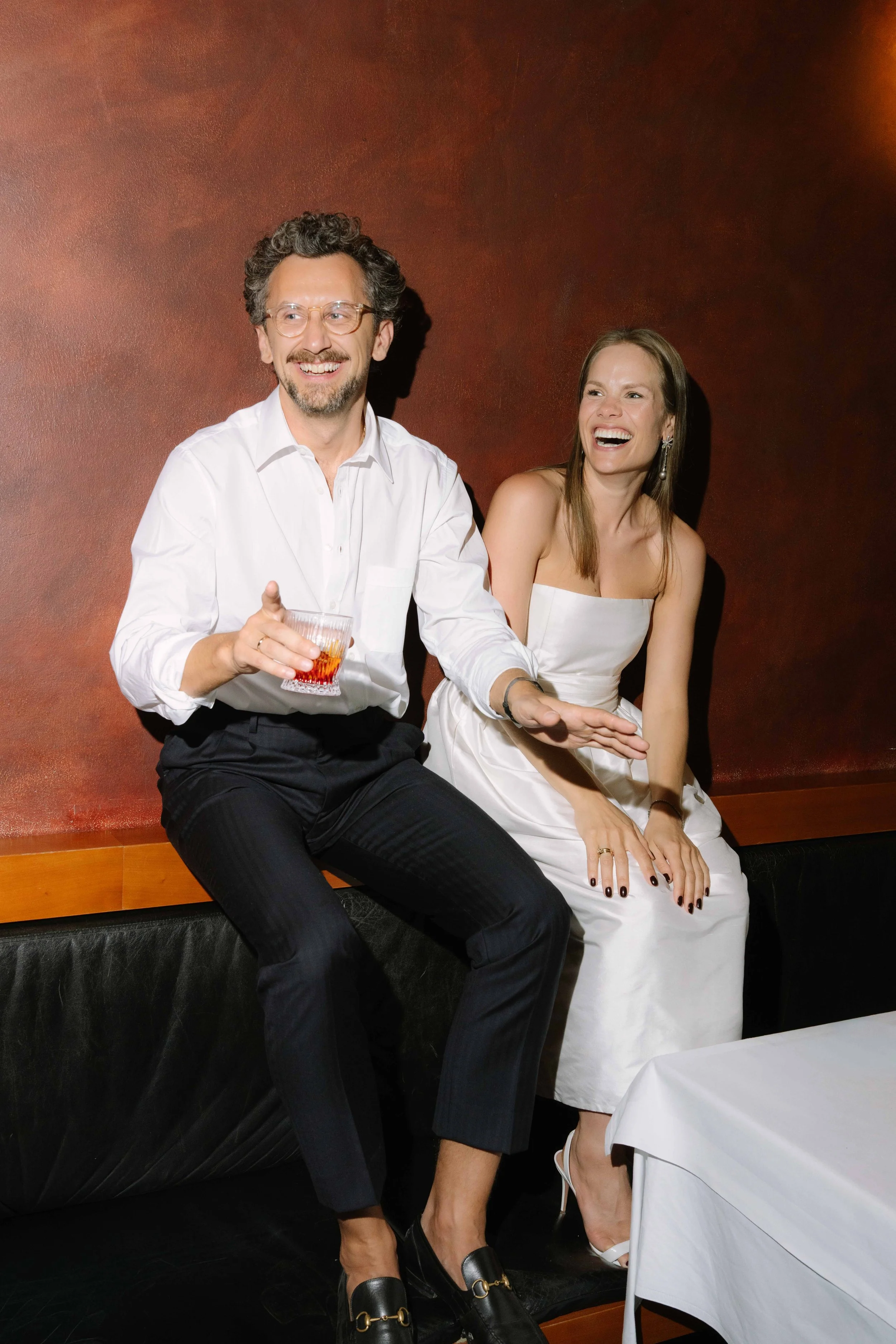 A man and woman sitting on a bench, laughing and enjoying drinks, dressed in formal attire at a social gathering.