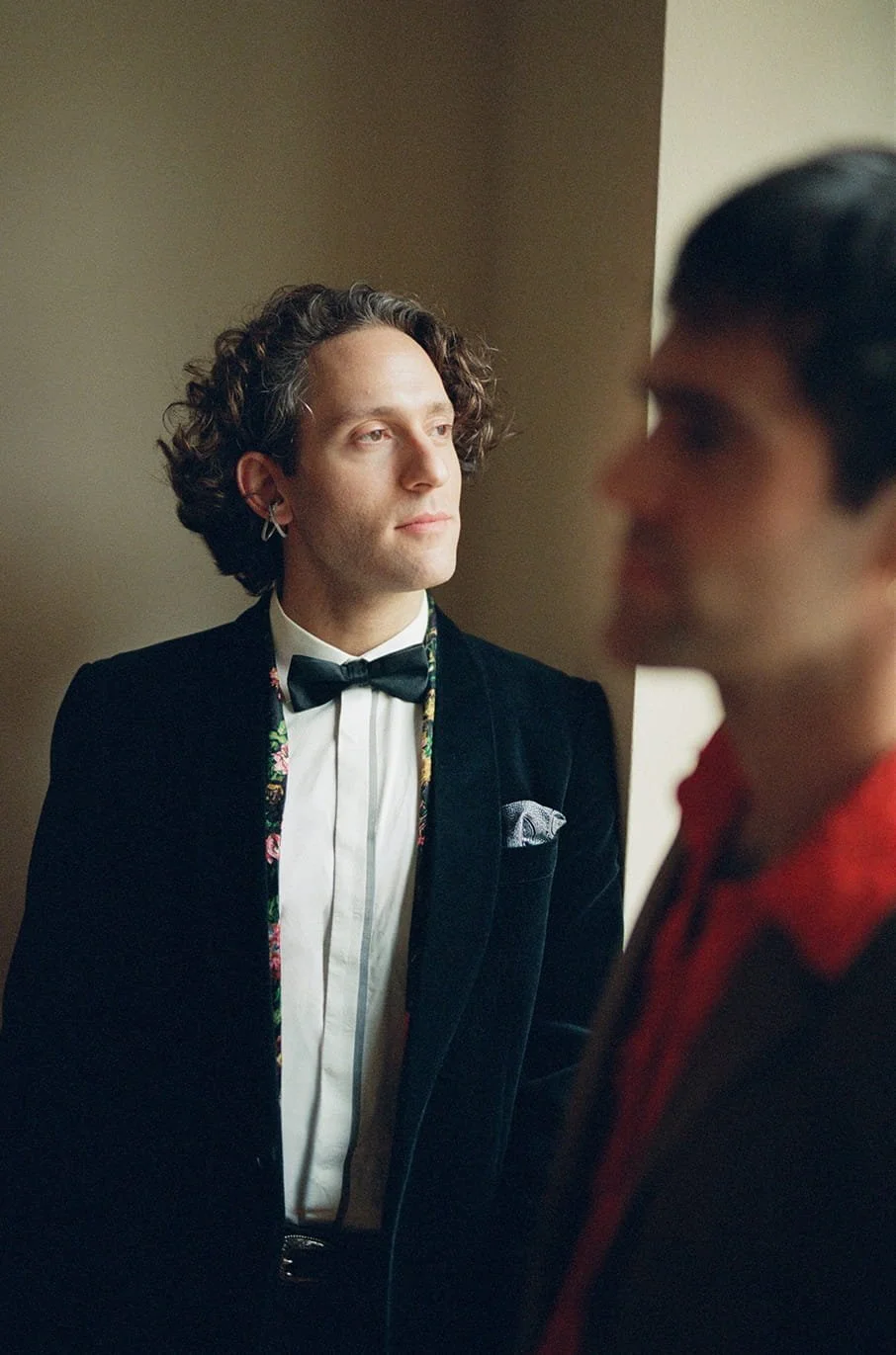 Two men in formal attire, one with curly hair and a black tuxedo, the other with short dark hair in a dark suit, standing close indoors near a beige wall.
